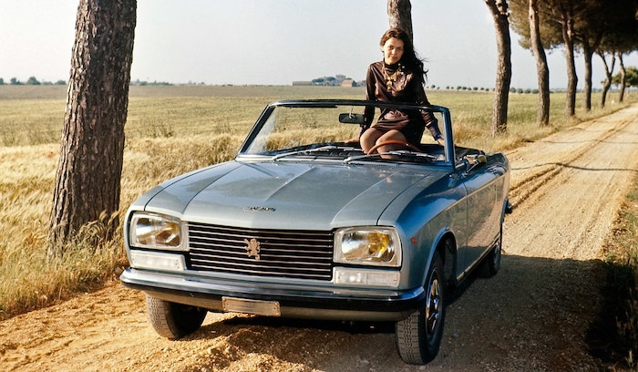 Peugeot 304 S grise de face avec une jeune femme à bord sur un chemin ensoleillé