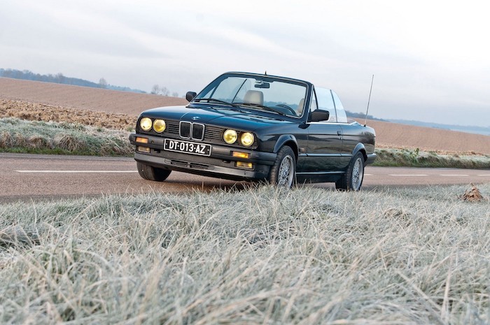 BMW E30 cabriolet de 3/4 face sur une route de campagne