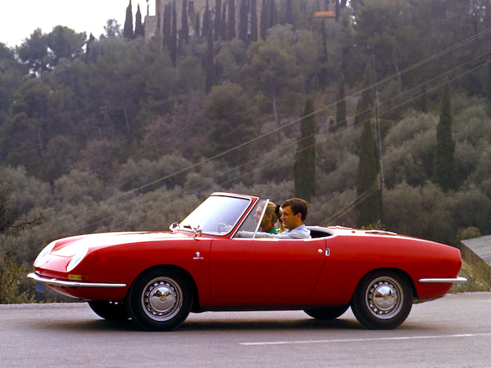 Fiat 850 Spider rouge avec un jeune couple à son bord sur une route italienne