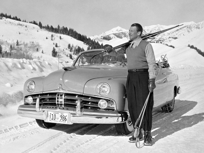 Homme tenant des skis devant un cabriolet Lincoln des années 50 de 3/4 face en montagne