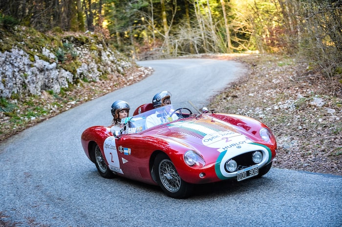 Osca 372 FS rouge au Tour Auto sur une route de forêt avec un couple à bord